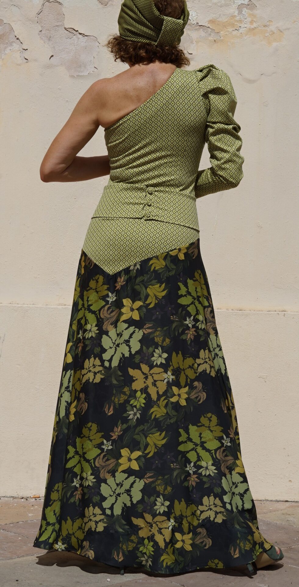 Vestido largo EMMA de EVA TYCHÉ en verde oliva, con escote asimétrico, un hombro descubierto, una manga larga, cuerpo de estampado geométrico y falda de estampado floral. / EMMA olive-green maxi dress by EVA TYCHÉ with an asymmetric neckline, one exposed shoulder, one long sleeve, a geometric-print bodice and a floral-print skirt.